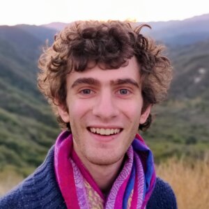 Photo of Samuel Breslow, a white man in his 20s wearing a blue sweater and purple scarf, smiling in the San Gabriel Mountains