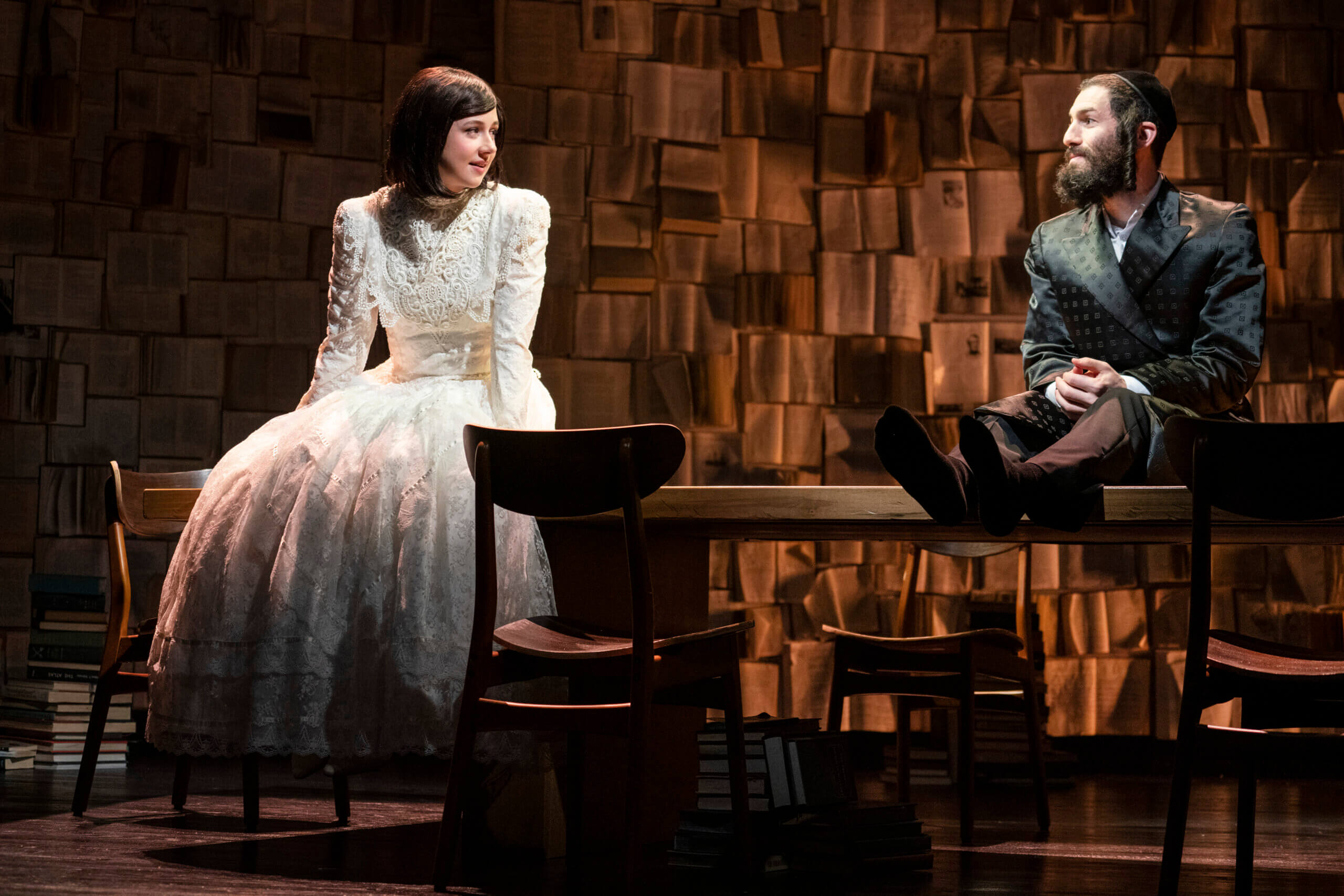 Two actors in Haredi attire sit on a wooden table, talking. 