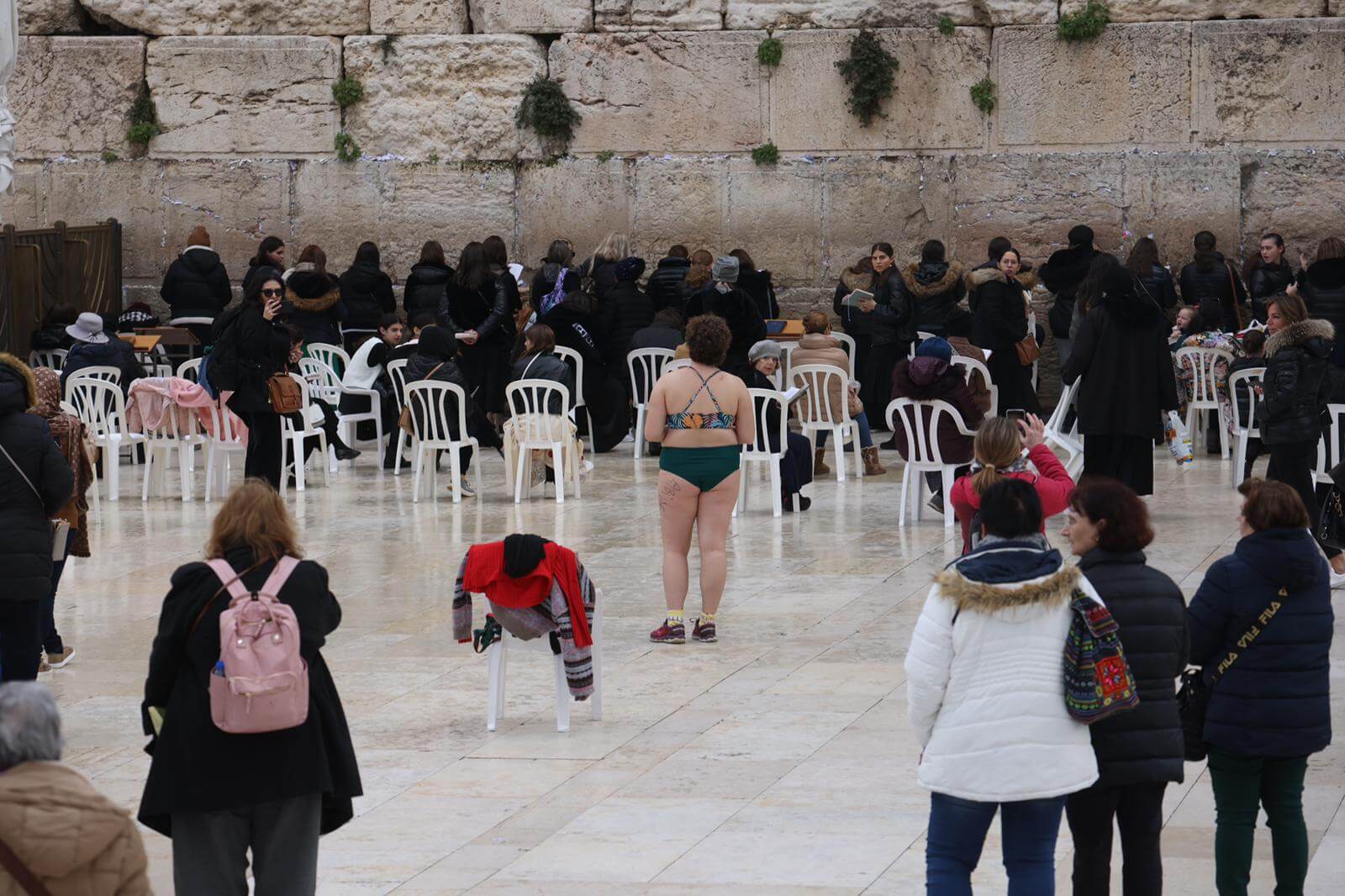 A woman stands in front of the Kotel in Jerusalem, her discarded clothing on the chair behind her.