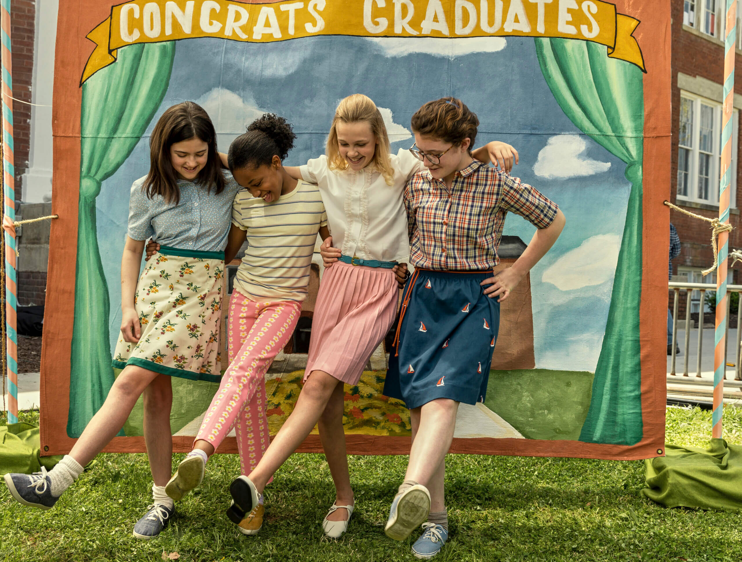 A still photo showing four preteen girls in 1970s attire dancing at a school event.