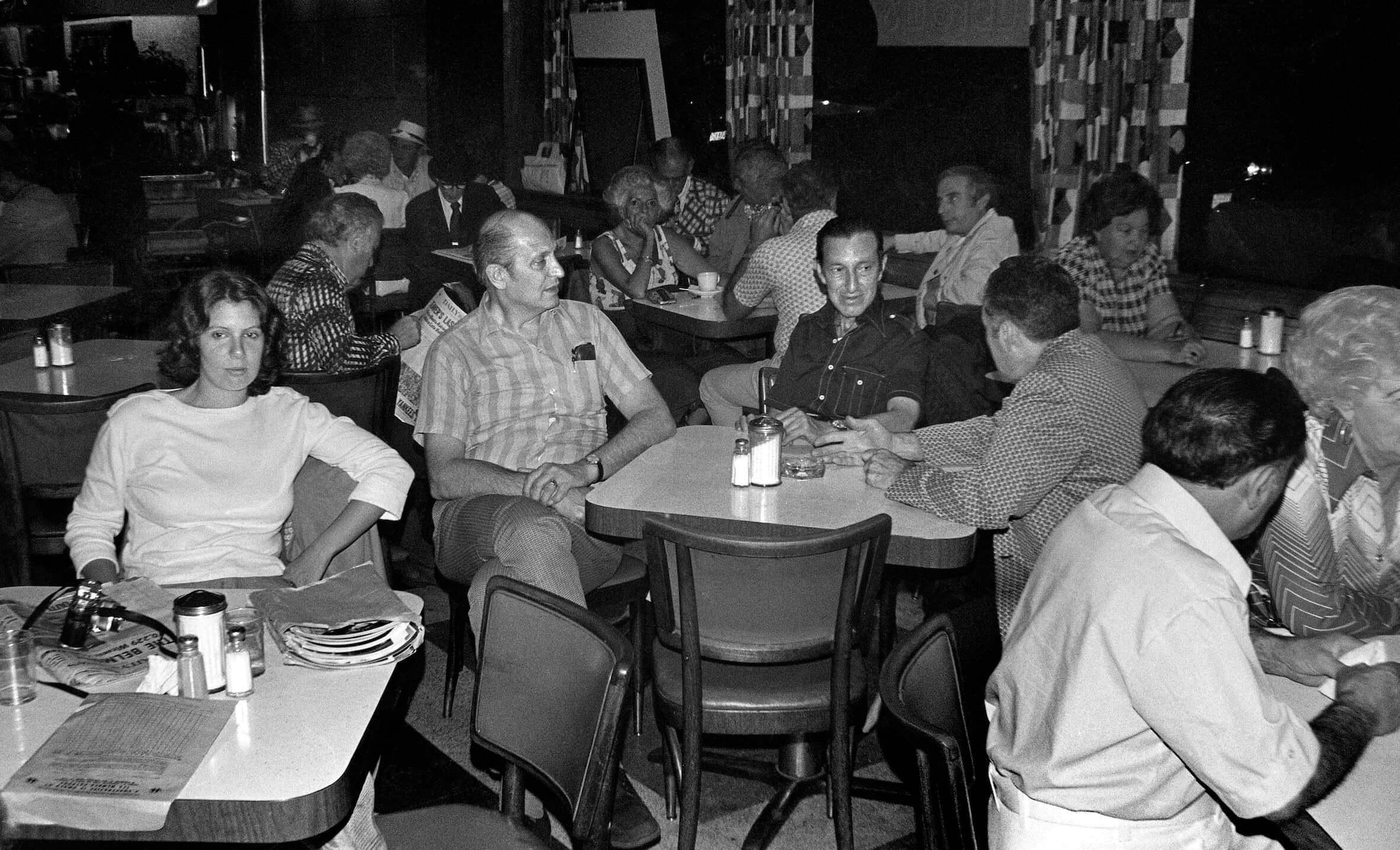 On the left, photographer Marcia Bricker Halperin sits at a table alone with an envelope full of photos. In the center, three men sit at another table conversing.