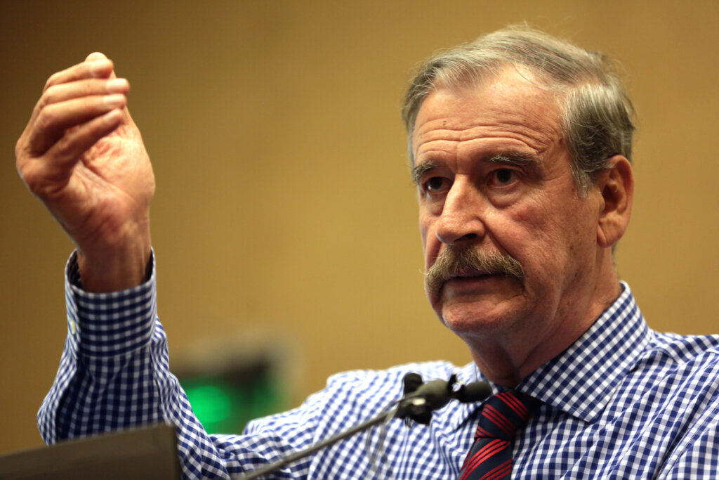 A middle-aged, White man with a light brown moustache and short hair, wearing a blue-and-white checkered shirt with a red tie, stands at a podium, his right hand up in the air.