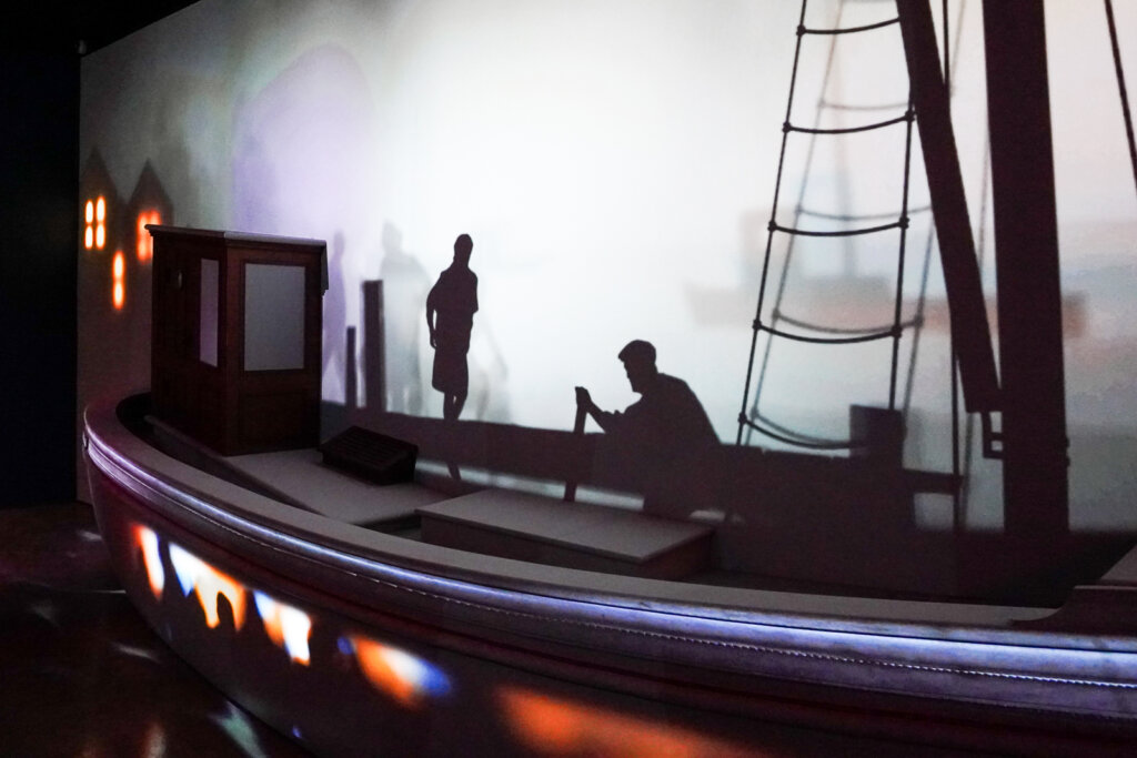 Model of a boat against a wall with two silhouette figures, one of a person standing up, one of a person rowing.