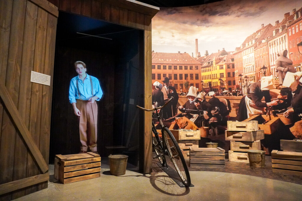 A boy in suspenders and brown pants stands talking, illuminated above a wooden structure. A bicycle leans against the structure. In the background, colorfully painted buildings and women with white bonnets working.