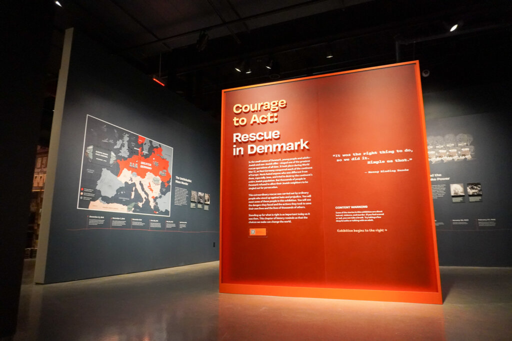 A red panel that says "Courage to Act: Rescue in Denmark" next to a wall with a gray map of Western and Central Europe, the Northern portion painted red with the words "Great Germany" on it.