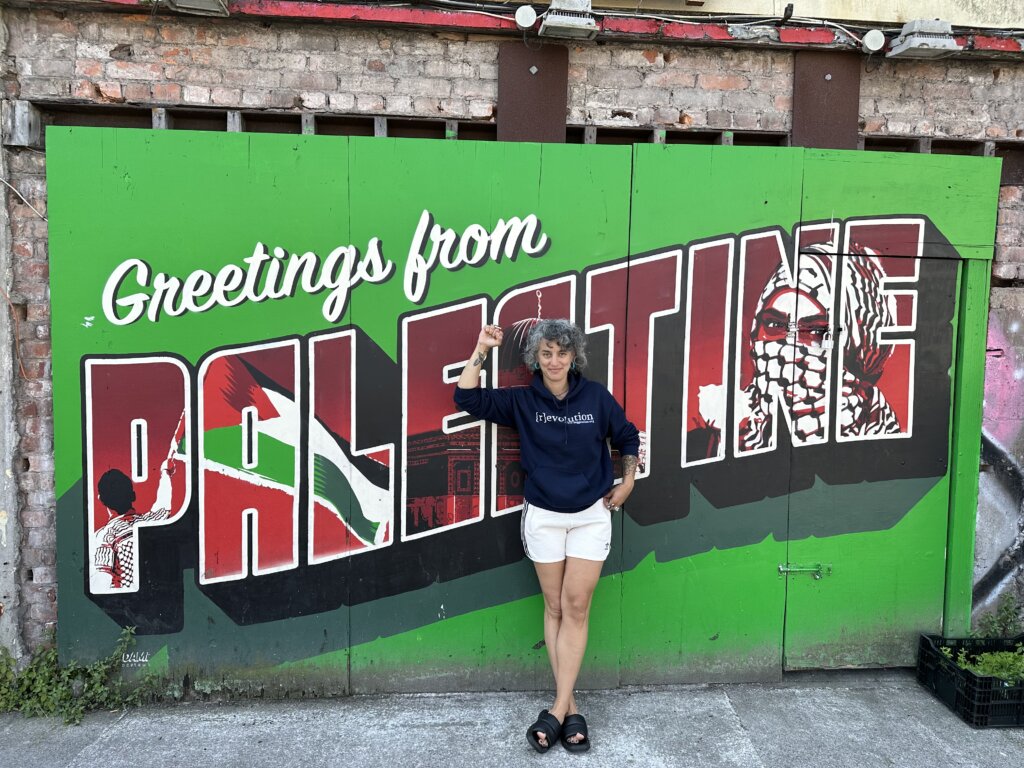 Woman stands holding up a raised fist in front of a green mural that says "Greetings from:" in script, then "PALESTINE" in red block letters with a person waving a Palestinian flag and a woman waving a kaffiyeh drawn into the letters.