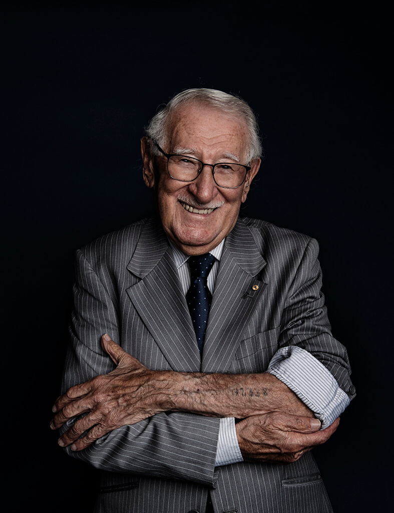 Man in pinstriped suit smiles broadly with sleeve rolled up to reveal number tattooed on forearm.