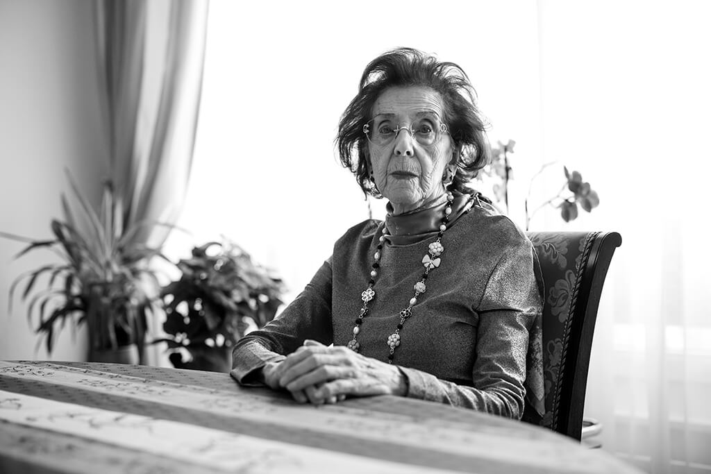 An older woman wearing a beaded necklace sits at a table with bright light and plants behind her, staring directly into the camera solemnly.