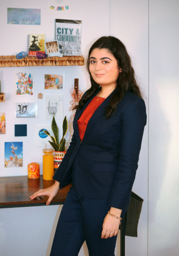 A young woman in a dark blue blazer and a red blouse smiles at the camera with one hand on a desk behind her. 