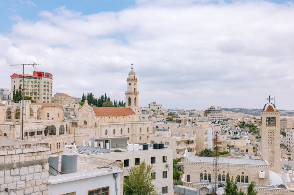 Landscape view of city with old tan-colored buildings, spires, red roofs, trees, clear sky. 