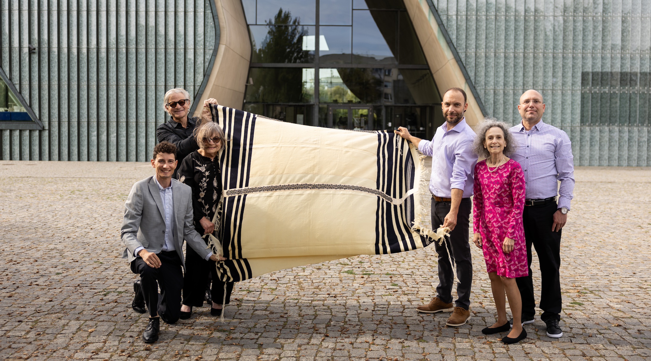 Lang family cousins hold Max Lang’s tallis in front of the POLIN Museum in Warsaw, on the day that they donated the prayer shawl to the museum. On the left, Stuart Schear (at rear), Jessie Lang and Jon Hanford. On the right, Theodore Hautin (holding the tallis), Annie Lang and Raphael Hautin. (Maciek Jazwiecki for POLIN Museum)