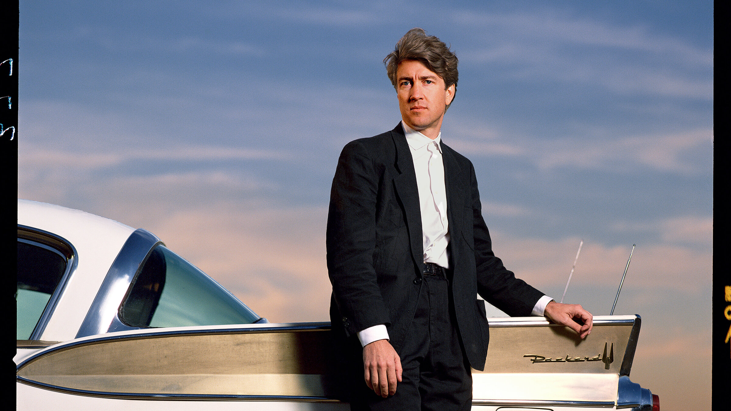 David Lynch stands outdoors in front of Packard vehicle in Los Angeles in 1986.